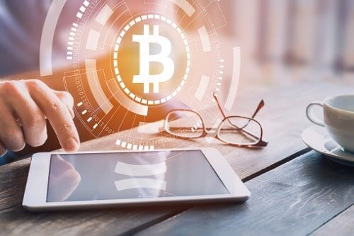 Person at a table using a tablet with a graphic of the bitcoin logo hovering above the tablet. Glasses and a cup are also sitting on the table